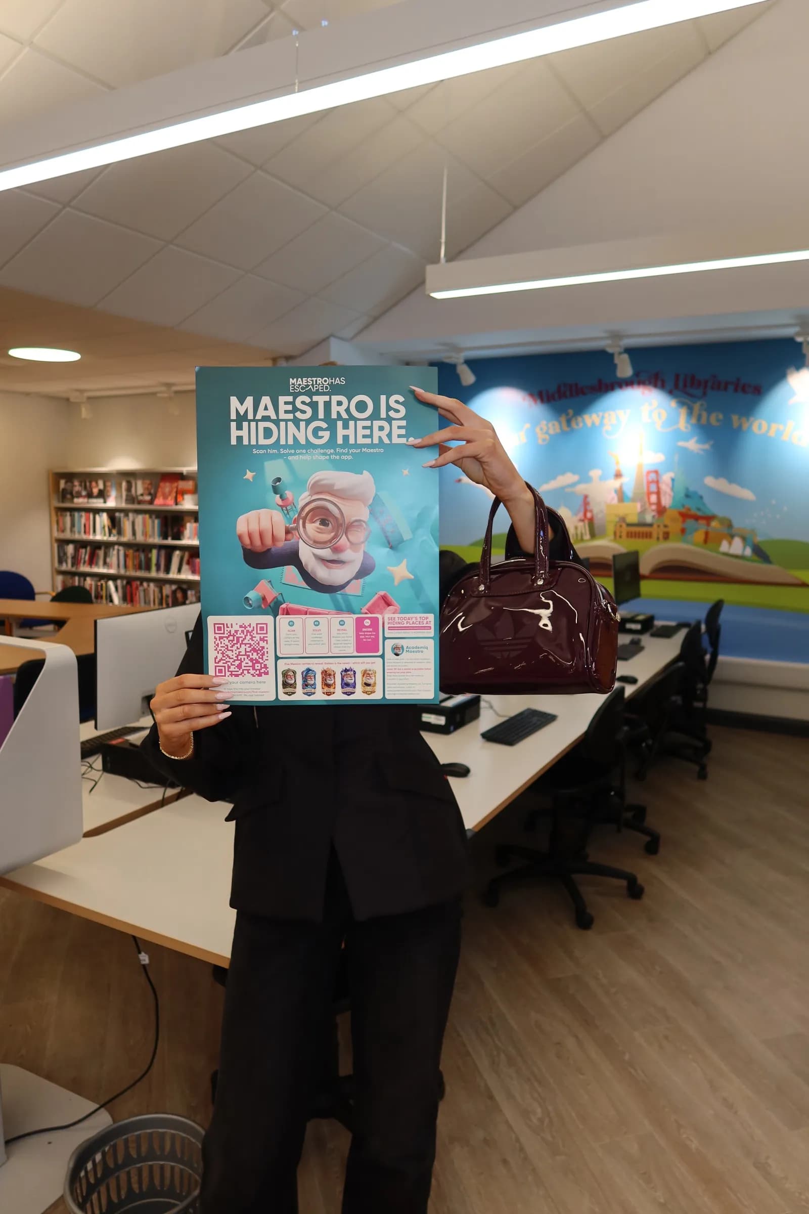 A person in a black suit holds a poster in front of their face that reads "Maestro is Hiding Here," featuring a cartoon character holding a magnifying glass.