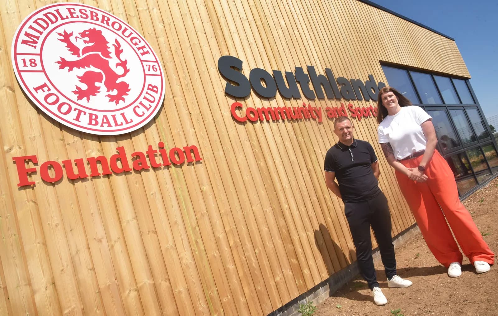 A man and a woman stand outside a building with wooden siding and a sign for Middlesbrough Football Club Foundation and Southlands Community and Sports.