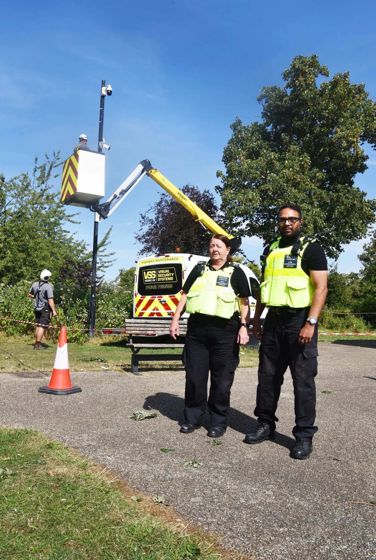 Two security guards in high-visibility vests stand in front of a white van with "Visual Security Systems" written on its side, next to a cherry picker hoisting a worker towards a pole with CCTV cameras.