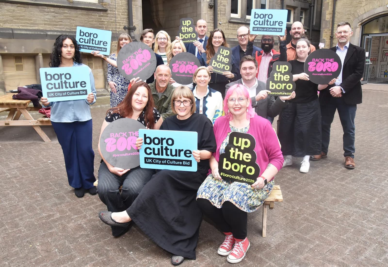 A diverse group of people, some seated and some standing, hold up various signs with phrases like "boro culture," "up the boro," and "BACK THE BORO." One woman with pink hair smiles at the camera while holding an "up the boro" sign.